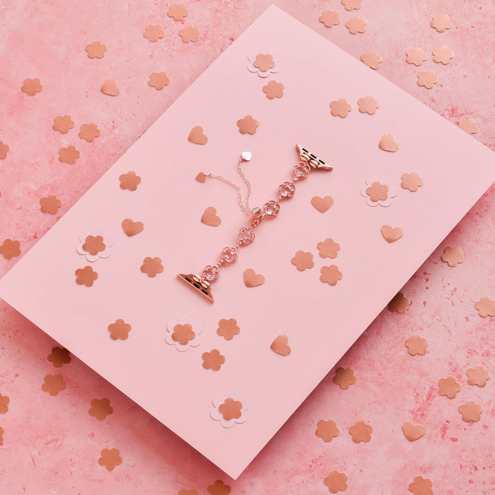 A delicate Rose Gold Flower Bracelet Apple Watch Strap by Coconut Lane, showcasing multiple pink beads and diamond-shaped charms, is laid on a pink card adorned with metallic flower and heart confetti. The background is a textured pink surface, adding charm and elegance, further enhanced by the sophisticated allure of the rose gold accessory from Coconut Lane.