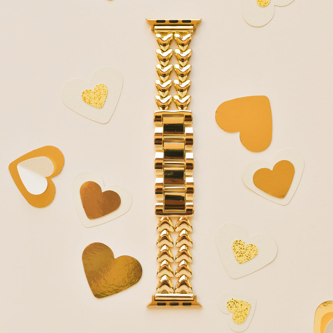 An intricately designed gold watch band is laid flat on a light-colored surface. Surrounding the Coconut Lane Hearts Chain Apple Watch Strap - Gold are assorted heart-shaped decorations in gold and white, with some featuring glitter accents. This elegant stainless steel adjustable strap fits various Apple Watch models.