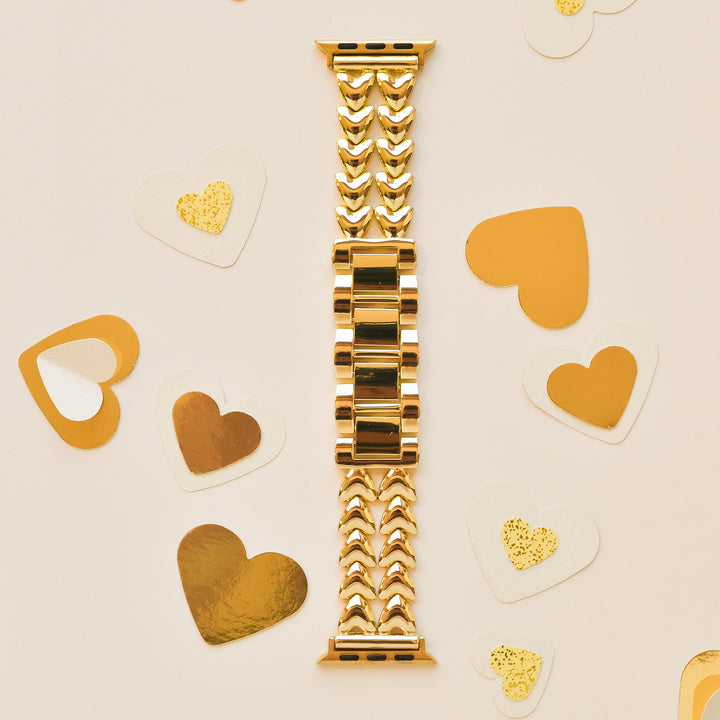 An intricately designed gold watch band is laid flat on a light-colored surface. Surrounding the Coconut Lane Hearts Chain Apple Watch Strap - Gold are assorted heart-shaped decorations in gold and white, with some featuring glitter accents. This elegant stainless steel adjustable strap fits various Apple Watch models.