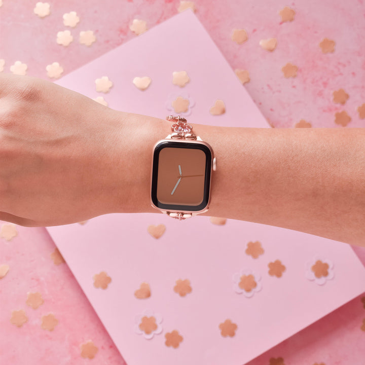 A person's wrist adorned with a stylish smartwatch, featuring the Rose Gold Flower Bracelet Apple Watch Strap from Coconut Lane. The background is a pink surface scattered with small heart-shaped and flower-shaped confetti, adding a festive touch.