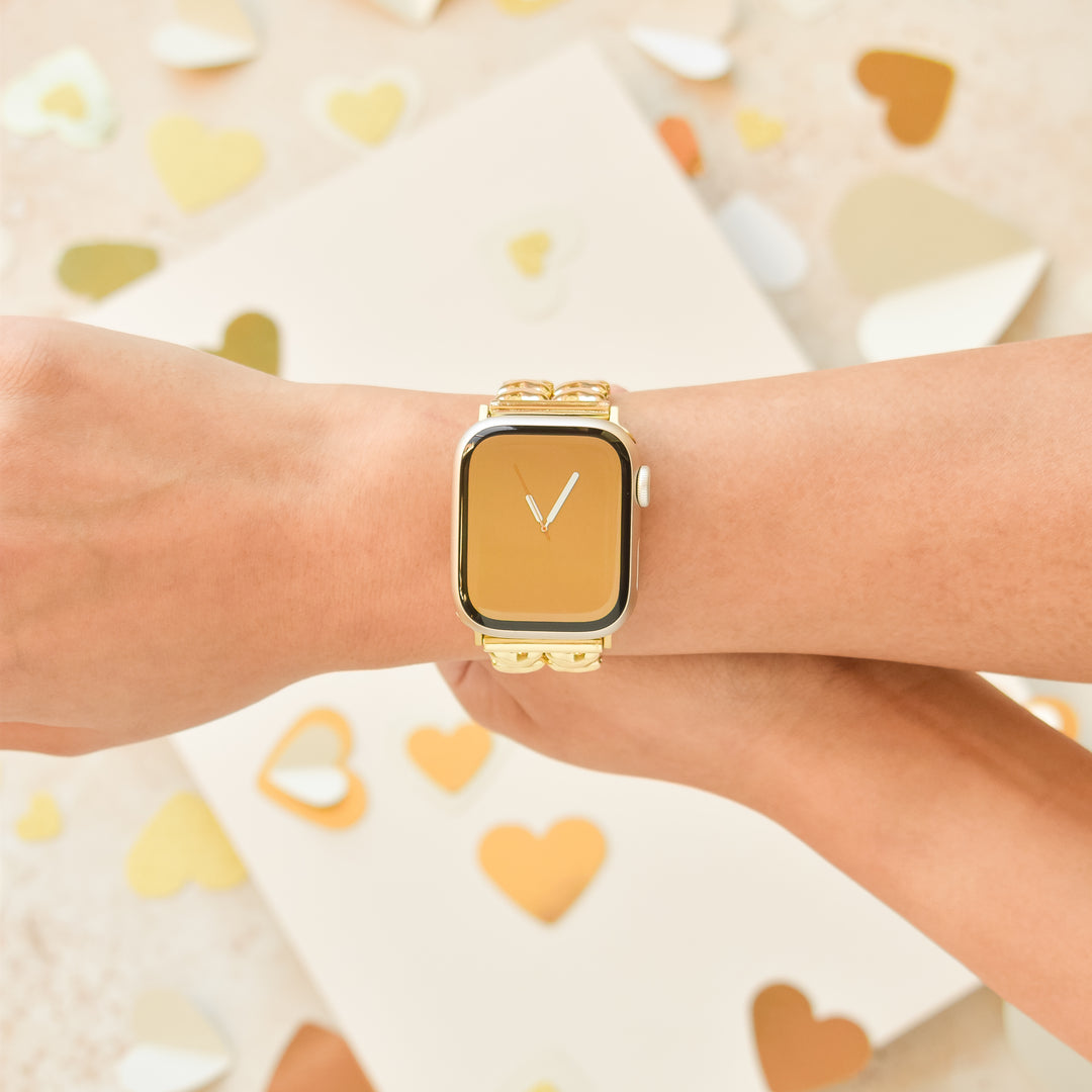 Close-up of a person wearing a gold smartwatch with the Coconut Lane Hearts Chain Apple Watch Strap - Gold. The background is adorned with various gold and cream-colored heart shapes. The person's hand is resting on what appears to be another arm or hand, elegantly showcasing the watch.
