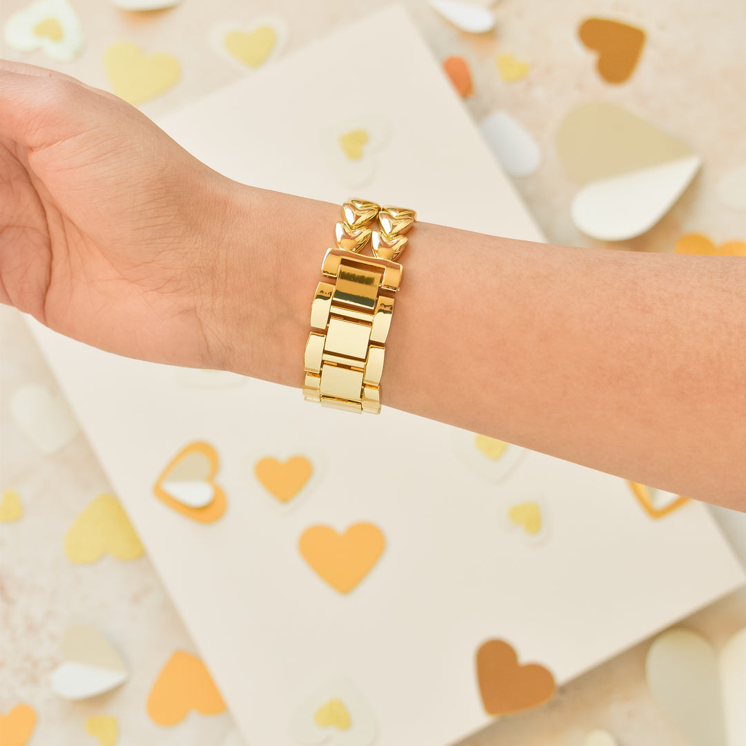 A hand adorned with Coconut Lane's Hearts Chain Apple Watch Strap - Gold is displayed against a backdrop sprinkled with gold, white, and beige heart-shaped confetti. The off-white surface in the scene adds a soft and elegant touch to the presentation.