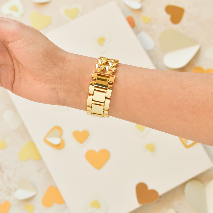 A hand adorned with Coconut Lane's Hearts Chain Apple Watch Strap - Gold is displayed against a backdrop sprinkled with gold, white, and beige heart-shaped confetti. The off-white surface in the scene adds a soft and elegant touch to the presentation.