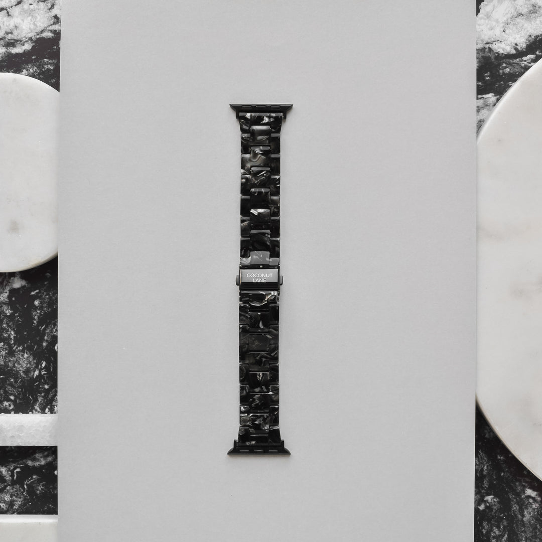 A Luxe Black Marble Apple Watch strap from Coconut Lane is displayed vertically against a light gray background. The band features a marbled pattern and a shiny metal clasp, with the brand name "Coconut Lane" engraved on it. The background includes marble accents in white and black on the sides.