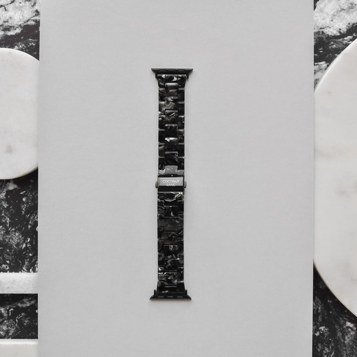 A Luxe Black Marble Apple Watch strap from Coconut Lane is displayed vertically against a light gray background. The band features a marbled pattern and a shiny metal clasp, with the brand name "Coconut Lane" engraved on it. The background includes marble accents in white and black on the sides.