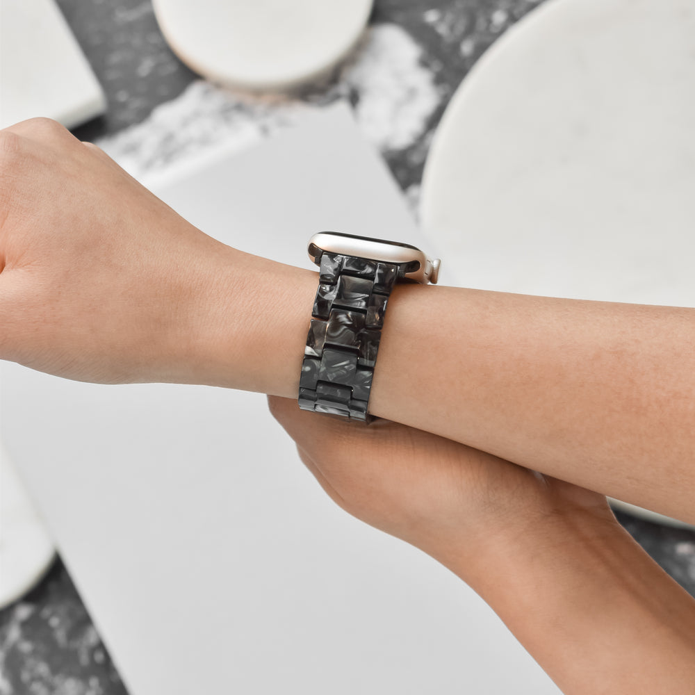 Close-up of a wrist adorned with a stylish smartwatch featuring Coconut Lane's Luxe Black Marble Apple Watch Strap. The strap boasts a glossy, marbled black texture, paired with a matte silver case. The background consists of a blurred, monochromatic surface with abstract black and white patterns, creating an overall modern and sleek aesthetic.