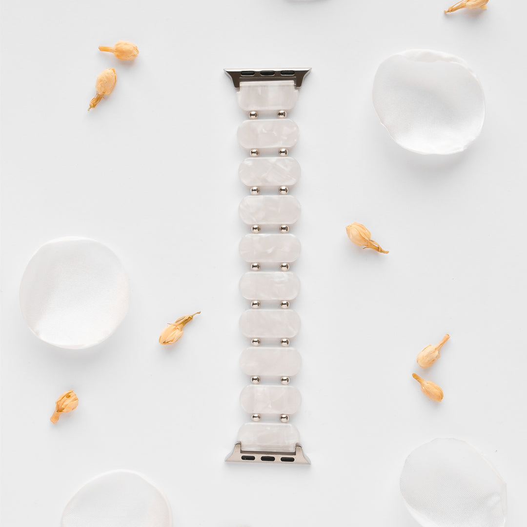 A White Pearl Beaded Apple Watch Strap by Coconut Lane, featuring a silver clasp, is positioned vertically in the center of a white background. Scattered white silk petals and small dried flowers surround it, creating an elegant and minimalist aesthetic. This unique Apple Watch accessory adds a touch of sophistication.