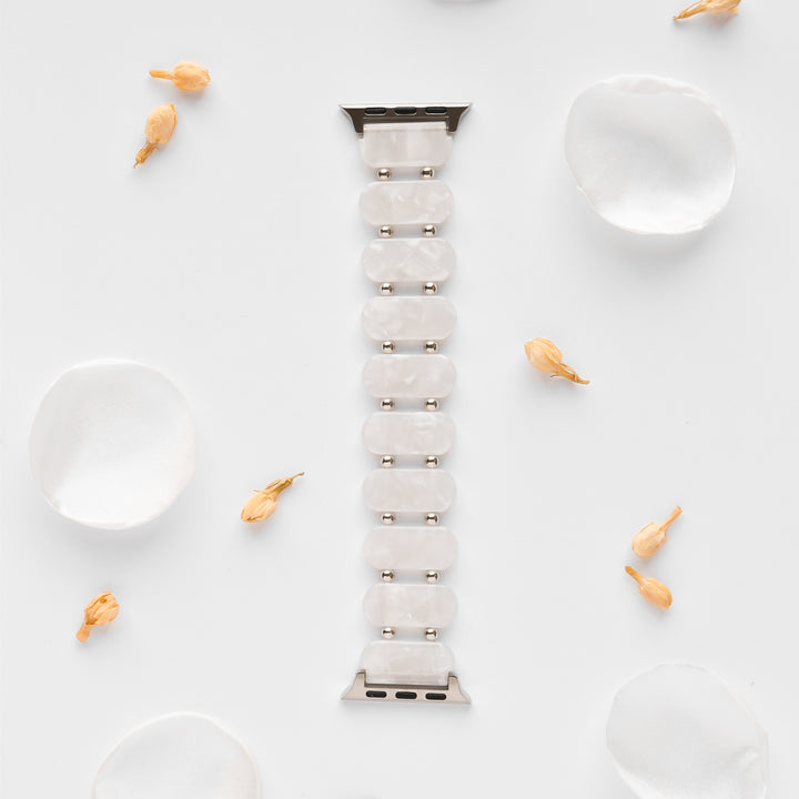 A White Pearl Beaded Apple Watch Strap by Coconut Lane, featuring a silver clasp, is positioned vertically in the center of a white background. Scattered white silk petals and small dried flowers surround it, creating an elegant and minimalist aesthetic. This unique Apple Watch accessory adds a touch of sophistication.