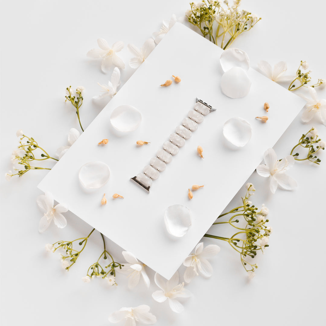 A stylish White Pearl Beaded Apple Watch Strap from Coconut Lane lies on a white rectangular board, surrounded by scattered white petals and small delicate white flowers. The setup is minimalist with a soft, elegant aesthetic, making it a unique Apple Watch accessory.