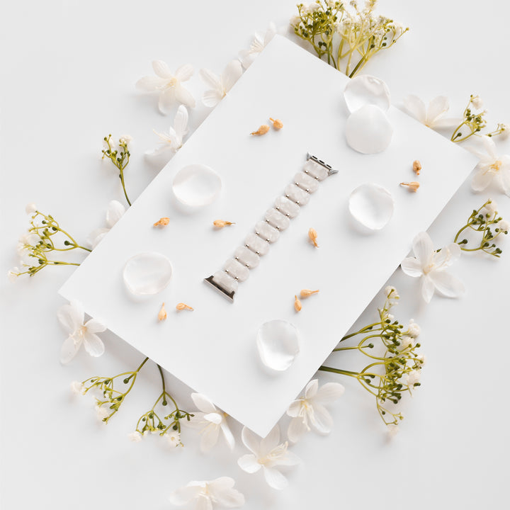 A stylish White Pearl Beaded Apple Watch Strap from Coconut Lane lies on a white rectangular board, surrounded by scattered white petals and small delicate white flowers. The setup is minimalist with a soft, elegant aesthetic, making it a unique Apple Watch accessory.