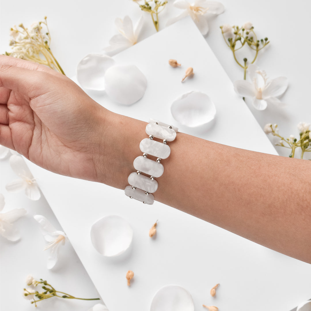 A person's arm showcasing Coconut Lane's White Pearl Beaded Apple Watch Strap, featuring elegant oval-shaped white stones, set against a white background. The scene is enhanced with white flowers and scattered petals, adding a delicate and refined touch.