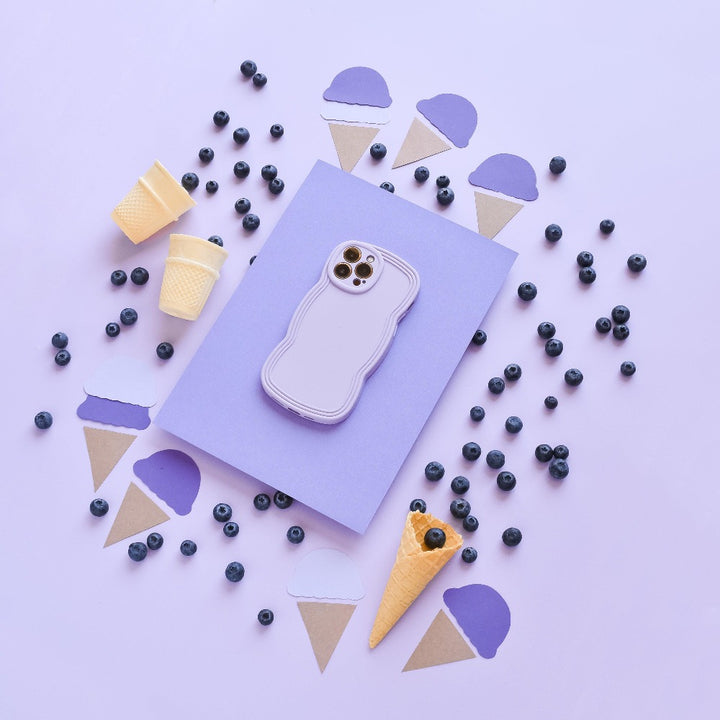 A pastel purple phone in a Coconut Lane NAKD Curvy Phone Case - Blueberry is placed on a light purple surface, surrounded by blueberries and cutouts of ice cream cones. Two small empty waffle cones and two yellow disposable cups also accompany the arrangement, all in a visually pleasing layout.