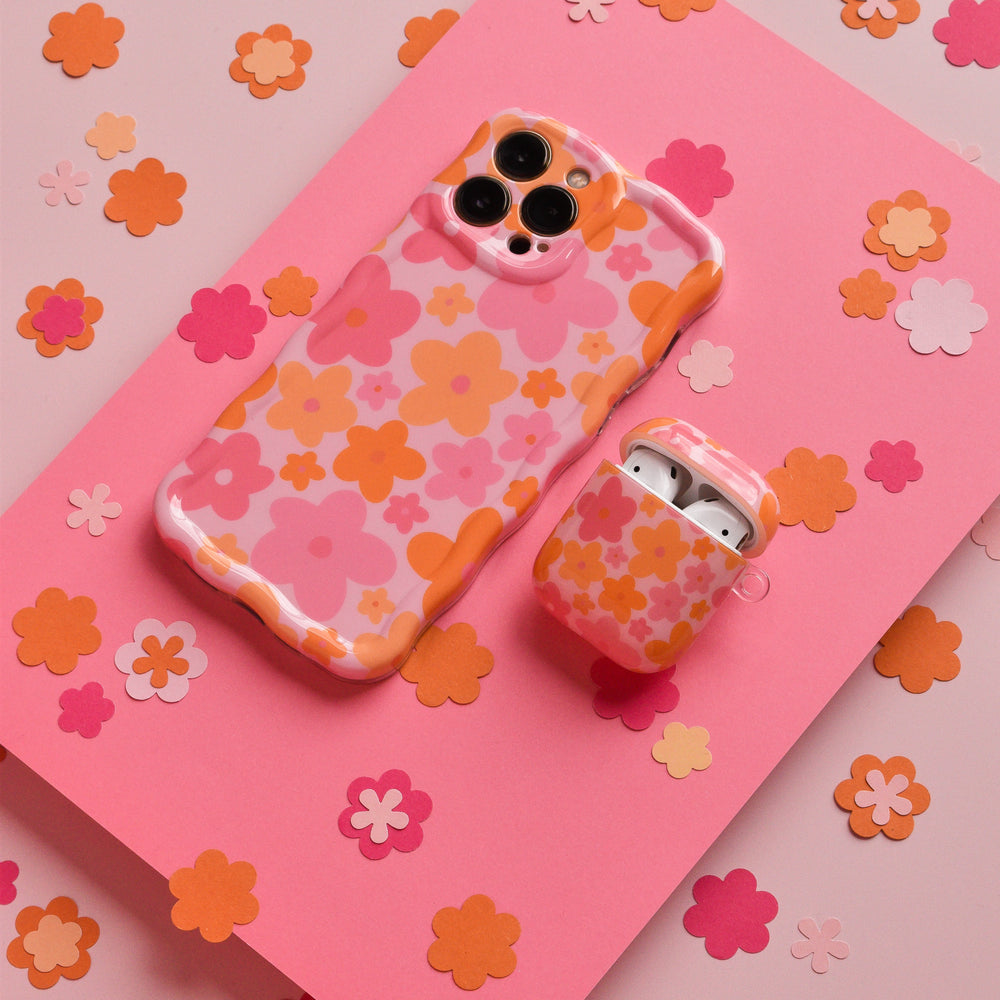 A smartphone and a pair of wireless earphones featuring the Coconut Lane Airpods Case - Pink & Orange Bloom with matching flower-patterned cases are placed on a pink background decorated with colorful flower cutouts. The AirPods case, adorned with pink and orange flowers on a white base, showcases an exquisite floral design crafted using IMD Print Technology.