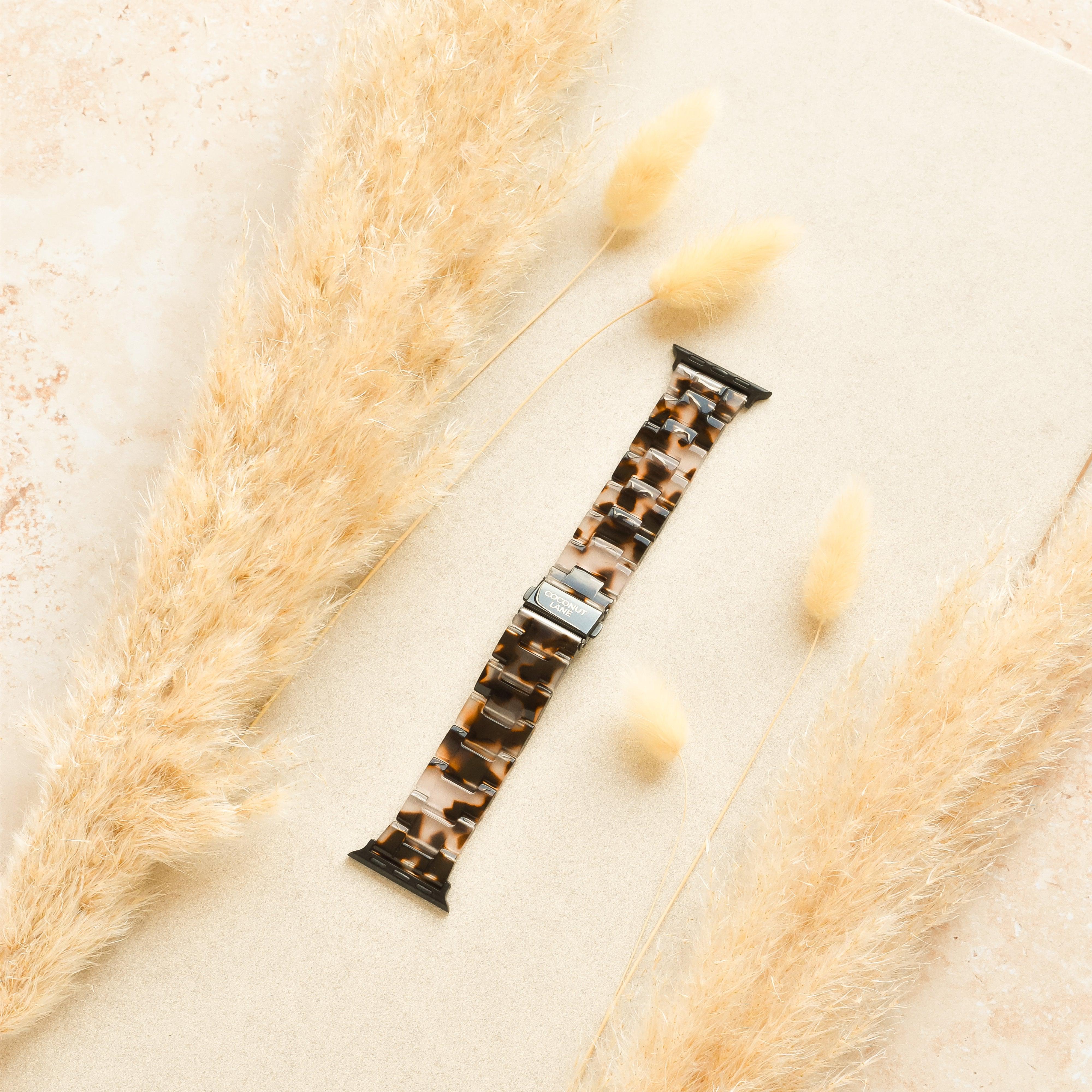 The Luxe Ivory Tort Apple Watch Strap by Coconut Lane, featuring a neutral beige resin with a tortoiseshell pattern, rests on a light-colored background surrounded by fluffy, beige dried pampas grass. The soft, natural textures create a warm, earthy aesthetic around the sleek adjustable strap.