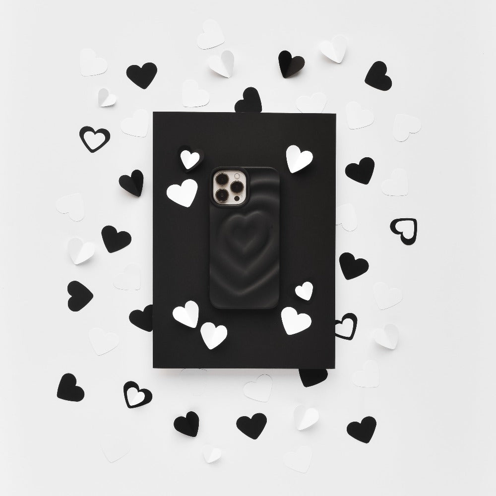 A Melting Heart Phone Case - Matte Black by Coconut Lane lies on a black rectangular backdrop. The backdrop is surrounded by scattered black and white paper hearts, creating a contrasting, monochromatic aesthetic on a white background.