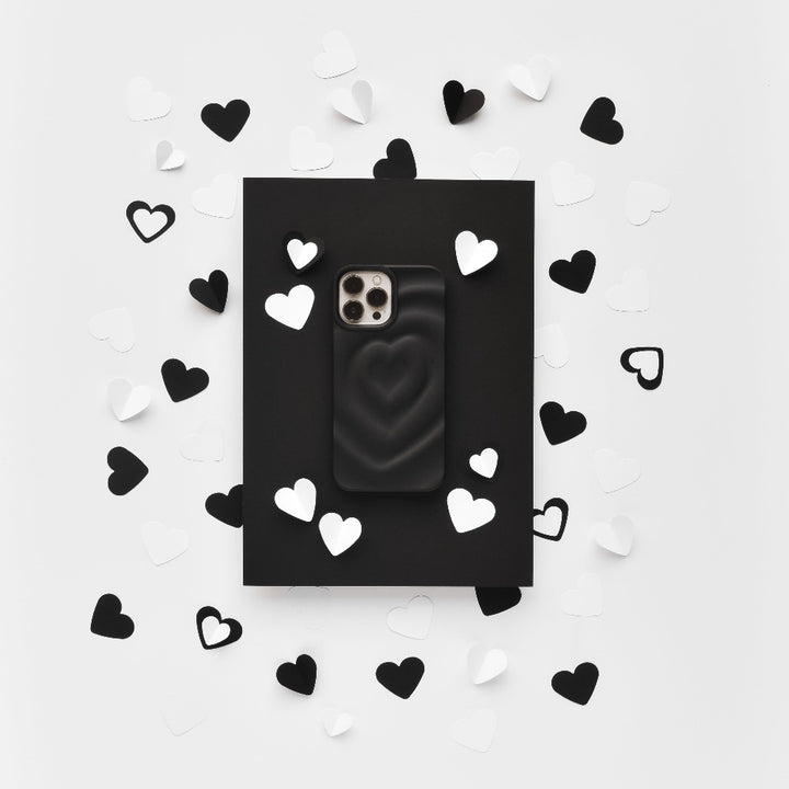 A Melting Heart Phone Case - Matte Black by Coconut Lane lies on a black rectangular backdrop. The backdrop is surrounded by scattered black and white paper hearts, creating a contrasting, monochromatic aesthetic on a white background.