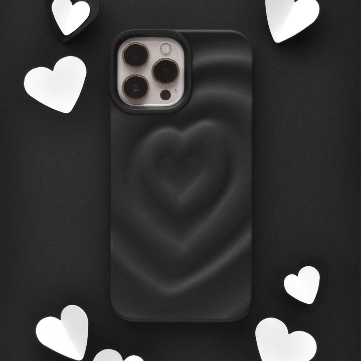 The Melting Heart Phone Case - Matte Black by Coconut Lane, featuring a 3D heart design, is displayed at the center of the image. Surrounding the phone case are several white and black heart-shaped cutouts, set against a dark background.