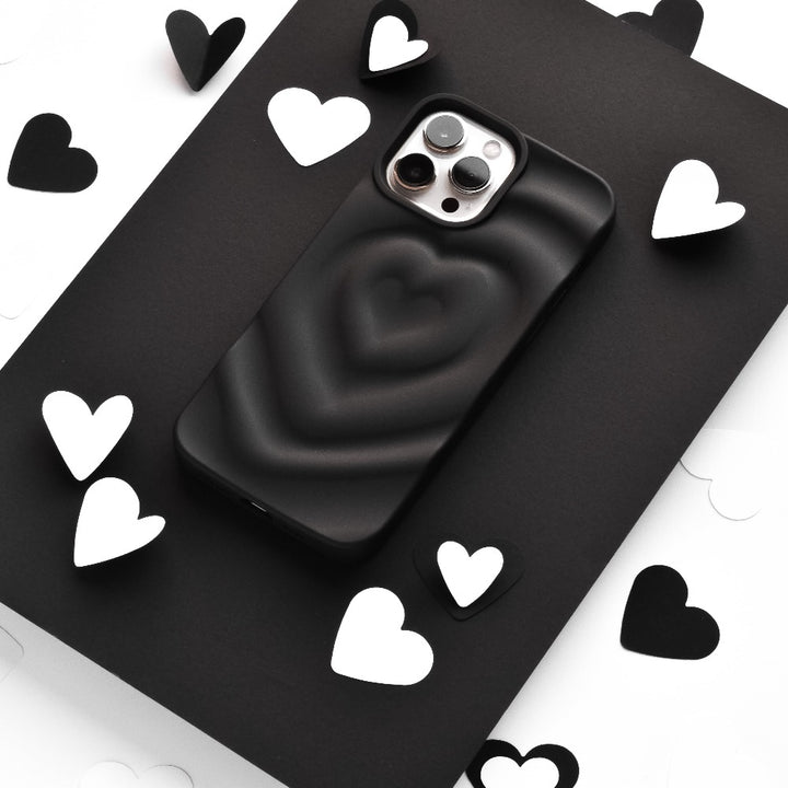 A smartphone encased in Coconut Lane's Melting Heart Phone Case - Matte Black sits on a black surface, surrounded by scattered black and white heart-shaped cutouts. The image emphasizes a monochromatic theme with a focus on the phone and the heart shapes.
