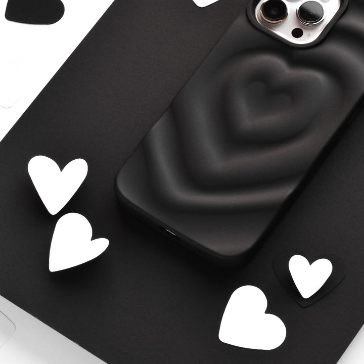 A black smartphone with the Coconut Lane Melting Heart Phone Case in Matte Black, featuring a raised heart design, is placed among scattered white and black heart-shaped cutouts on a flat surface. The phone's dual camera lenses are visible at the top.