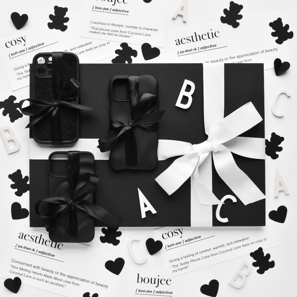 A black and white display of gift-wrapped items, potentially including protective 3D Boujee Phone Cases in a sleek matte black finish from Coconut Lane, adorned with bows. The arrangement is surrounded by decorative cutouts of hearts, letters, and teddy bears. The background features printed definitions like "cosy" and "aesthetic.
