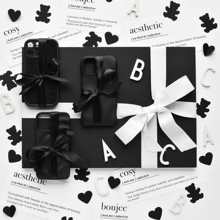 A black and white display of gift-wrapped items, potentially including protective 3D Boujee Phone Cases in a sleek matte black finish from Coconut Lane, adorned with bows. The arrangement is surrounded by decorative cutouts of hearts, letters, and teddy bears. The background features printed definitions like "cosy" and "aesthetic.
