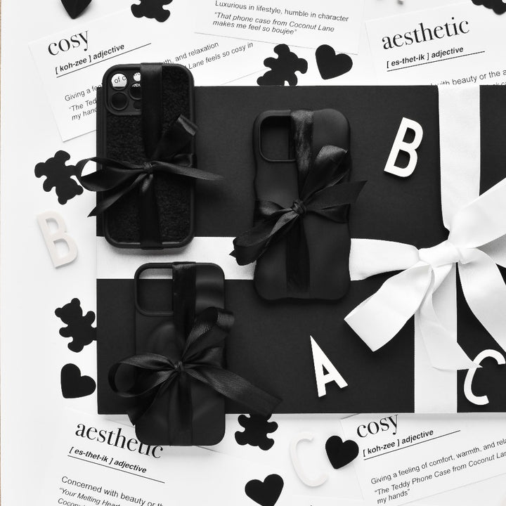 A black-and-white flat lay showcasing four Melting Heart Phone Cases - Matte Black by Coconut Lane, wrapped with black ribbons on a black background. Scattered around are cut-out letters, black paper hearts, and printed definitions of "aesthetic" and "cozy." A white ribbon bow is visible at the edge, enhancing the overall chic vibe.