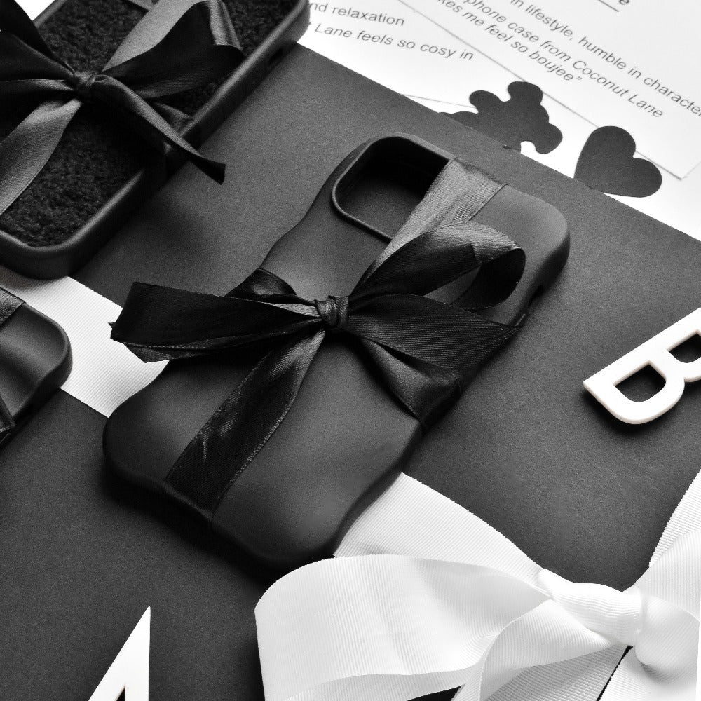 A flatlay image featuring neatly wrapped black gift boxes adorned with black and white ribbons. The central focus is on Coconut Lane's 3D Boujee Phone Case - Matte Black, elegantly tied with a black ribbon. The background showcases black and white text along with decorative elements in monochrome tones, all boasting a matte black finish.