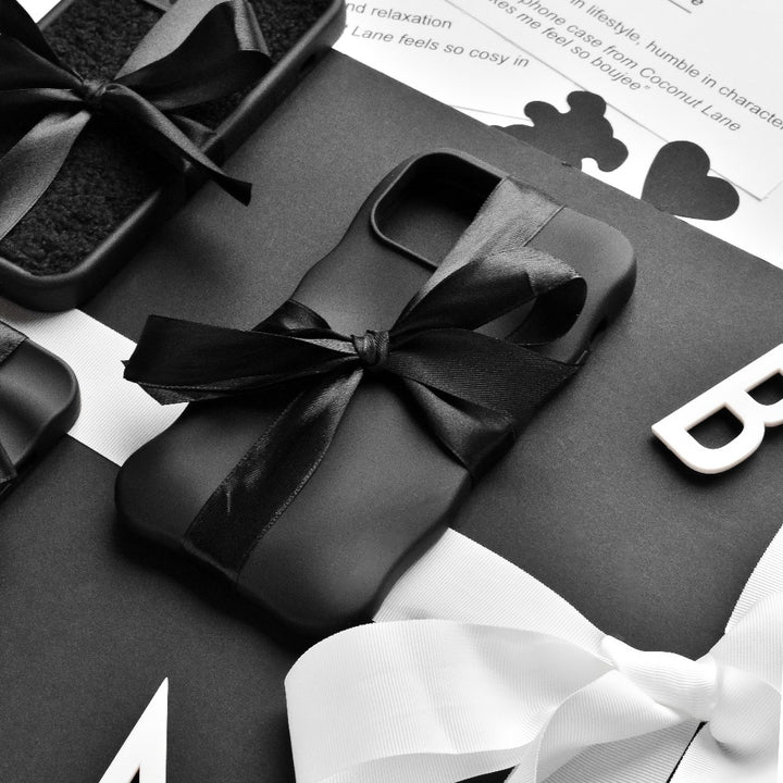 A flatlay image featuring neatly wrapped black gift boxes adorned with black and white ribbons. The central focus is on Coconut Lane's 3D Boujee Phone Case - Matte Black, elegantly tied with a black ribbon. The background showcases black and white text along with decorative elements in monochrome tones, all boasting a matte black finish.