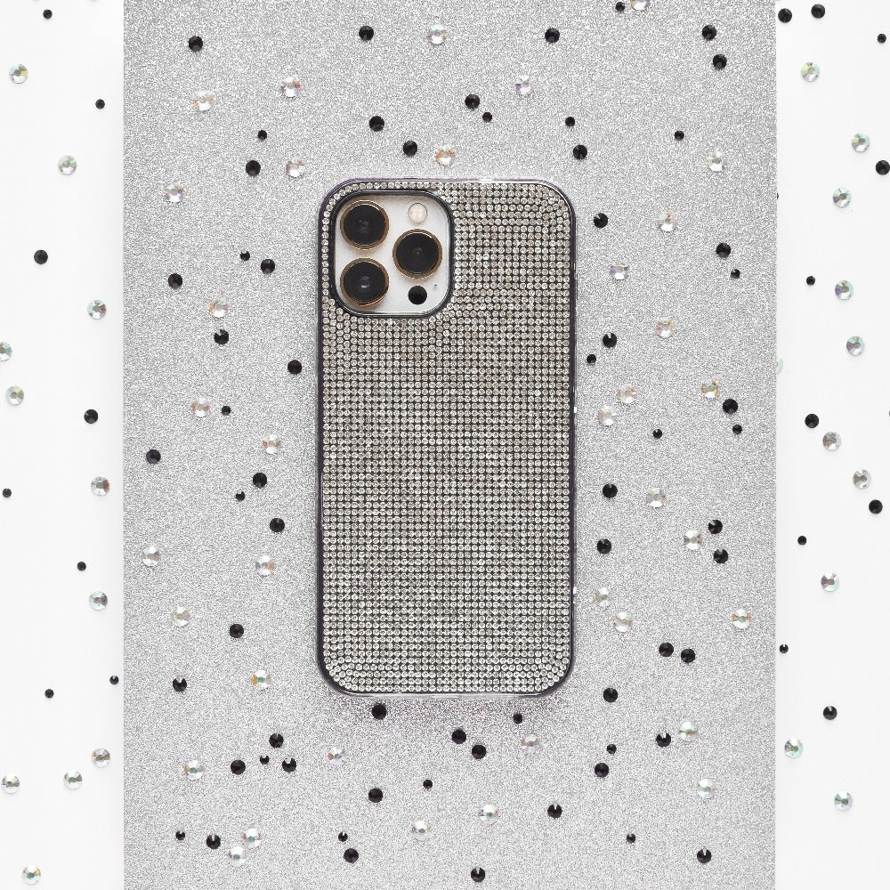 Against a sparkling silver background adorned with small black and white sequins scattered around, a smartphone is encased in the glittery Disco Sparkle Phone Case - Silver by Coconut Lane. The case features a textured, grid-like pattern and showcases three visible camera lenses on the back.