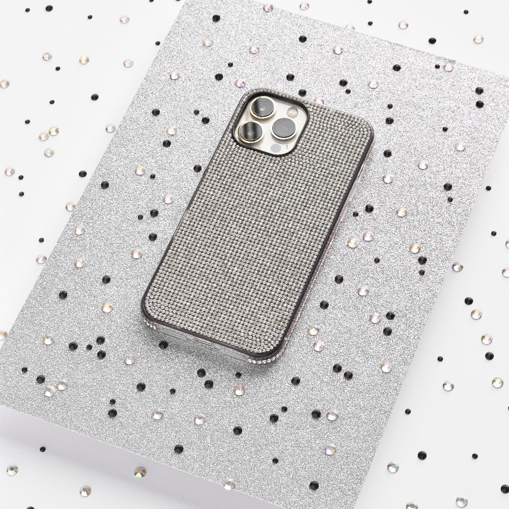 A silver iPhone adorned with the Coconut Lane Disco Sparkle Phone Case - Silver, showcasing a grid pattern, rests on a glittery silver surface. The background is sprinkled with small black and silver sequins, setting a festive and glamorous scene.