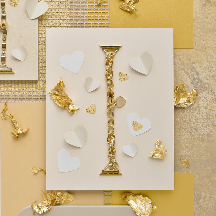 A flat lay of the Chain Bracelet Apple Watch Strap - Gold from Coconut Lane on a white card surrounded by small white hearts and gold foil pieces. The background features different shades of gold and beige paper textures, creating a luxurious and elegant aesthetic reminiscent of its design for various Apple Watch models.