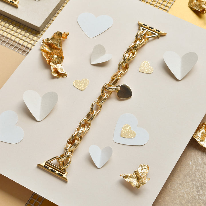 The Coconut Lane Chain Bracelet Apple Watch Strap in gold, featuring heart-shaped decorations, is elegantly displayed on a beige and white background. Gold leaf pieces and heart-shaped cutouts in white and glittery gold are scattered around, enhancing the romantic composition of this stainless steel metal adjustable chain bracelet.