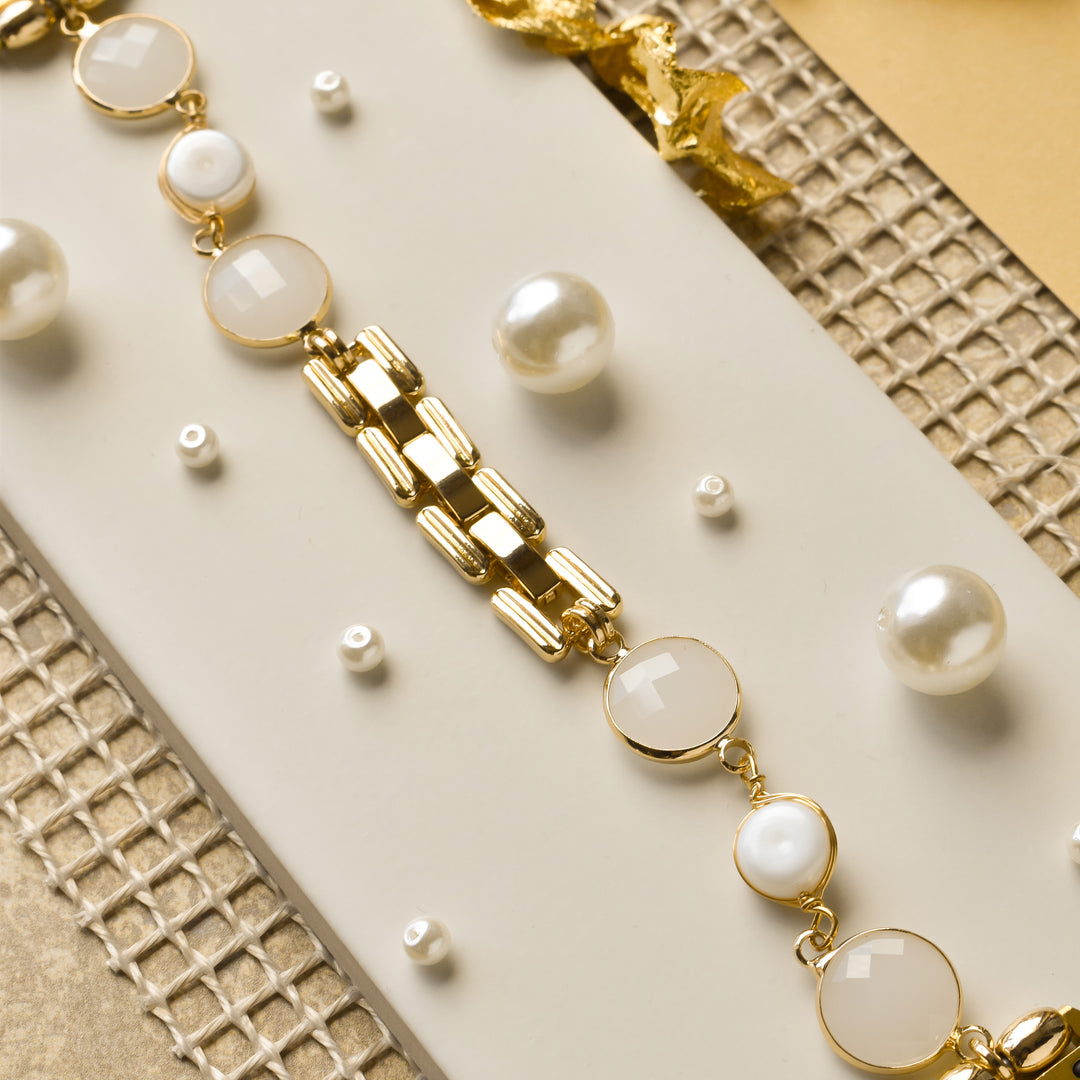 A close-up image of the Cute Stone Bracelet Apple Watch Strap by Coconut Lane placed on a beige background with a textured gold mesh underneath. The strap features alternating round pearl and faceted gold settings, complemented by scattered small pearls and a stainless steel metal ribbon accent nearby.