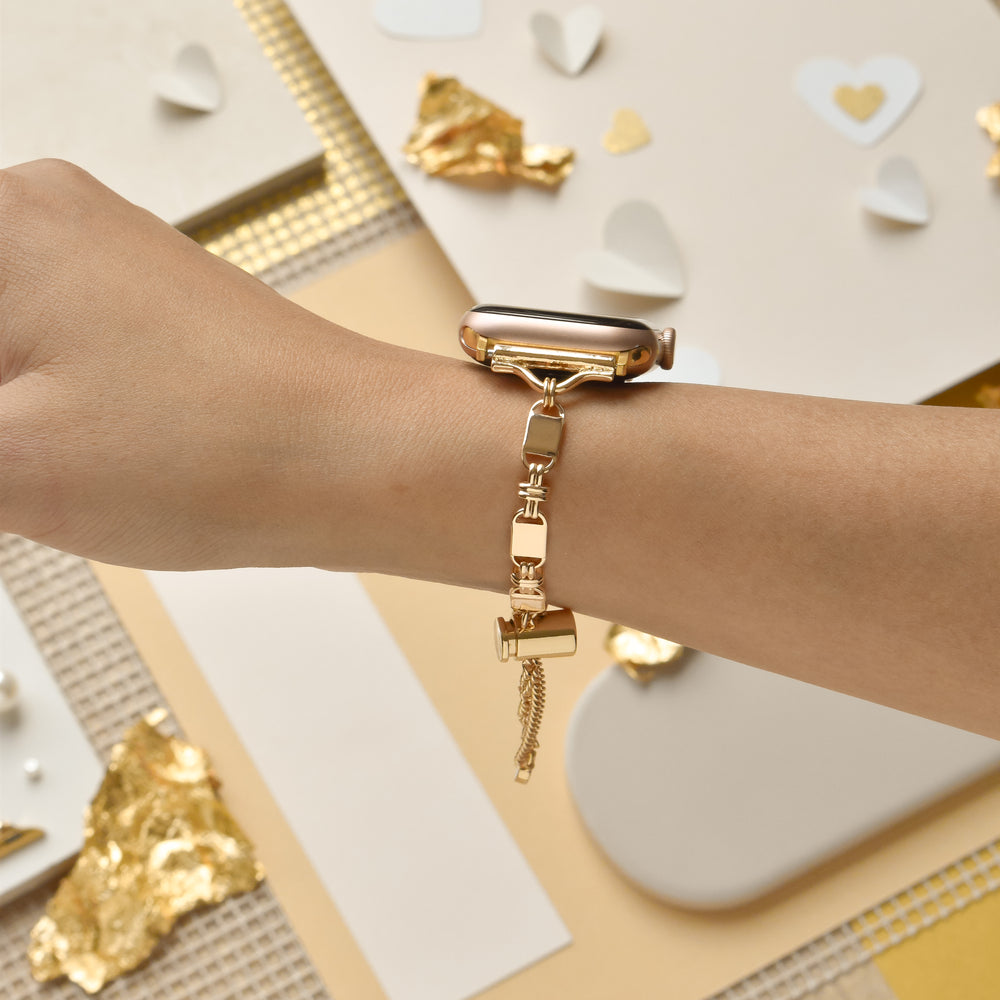A close-up of a person's arm adorned with Coconut Lane's Simple Gold Bracelet Apple Watch Strap, paired with a matching simple gold bracelet. The background features cream and gold hearts, gold foil, and textured materials, creating a stylish and elegant setting.