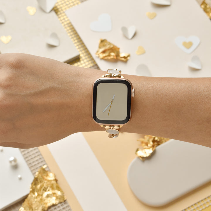 A person's wrist wearing a gold smartwatch with a simple clock face is shown, beautifully complemented by the Cute Stone Bracelet Apple Watch Strap from Coconut Lane. The background features various decorative items such as white paper hearts, gold foil pieces, and textured fabric, creating an elegant and festive atmosphere.