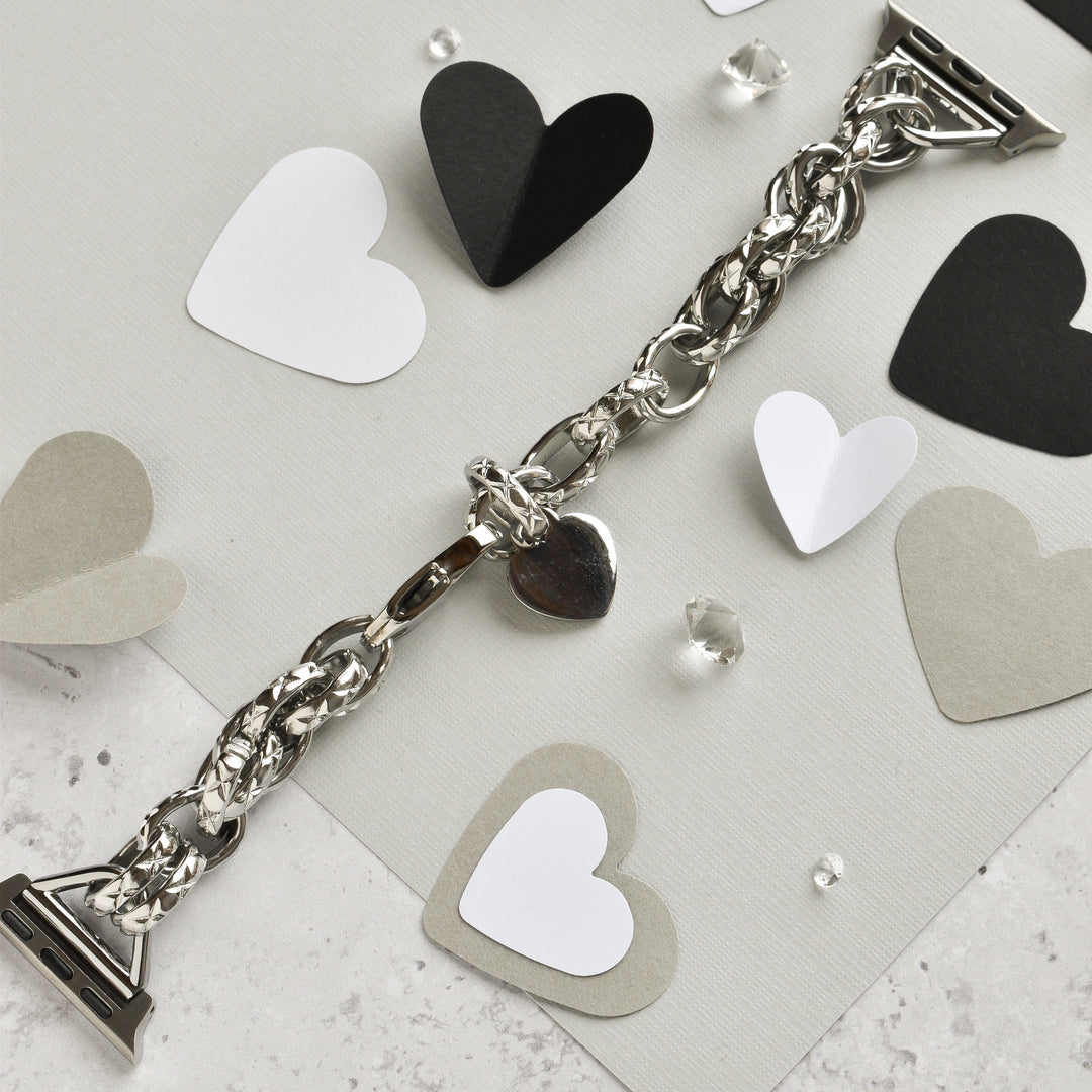 A Chain Bracelet Apple Watch Strap in silver, by Coconut Lane, rests on a textured surface adorned with black, white, gray, and silver paper hearts. Small transparent crystals are scattered around, adding a touch of sparkle to the composition. The scene is reminiscent of an elegantly crafted bracelet with a heart charm.