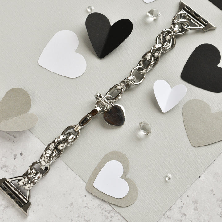 A Chain Bracelet Apple Watch Strap in silver, by Coconut Lane, rests on a textured surface adorned with black, white, gray, and silver paper hearts. Small transparent crystals are scattered around, adding a touch of sparkle to the composition. The scene is reminiscent of an elegantly crafted bracelet with a heart charm.