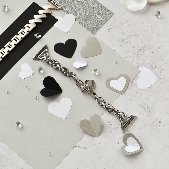 A Chain Bracelet Apple Watch Strap in silver by Coconut Lane lies on a textured surface surrounded by black, white, and gray paper hearts. Small clear gems are scattered around, adding a touch of sparkle to the arrangement. A cream-colored bracelet is visible at the top left of the image.