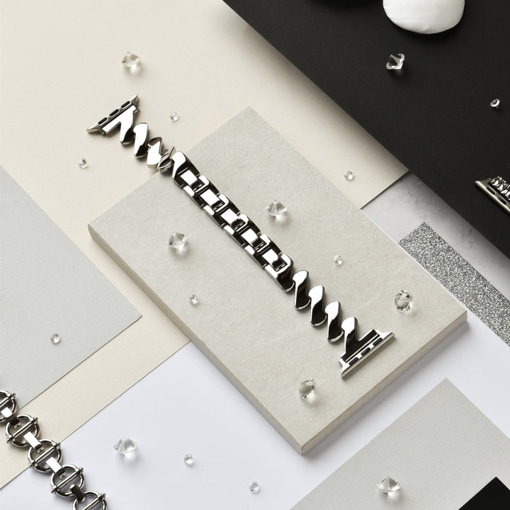 A Coconut Lane Get Wavy Apple Watch Strap - Silver with a sleek, interlocking design is displayed on a beige rectangular surface. Scattered around it are small, clear, gem-like decorations on a neutral background of black, white, and gray surfaces.