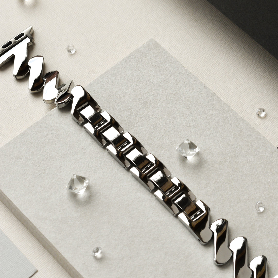 A close-up view of Coconut Lane's Get Wavy Apple Watch Strap - Silver, featuring a polished stainless steel band with a rectangular link pattern, displayed on a light gray backdrop. Small, clear crystals are scattered around the Apple Watch strap, adding a touch of elegance to the composition.