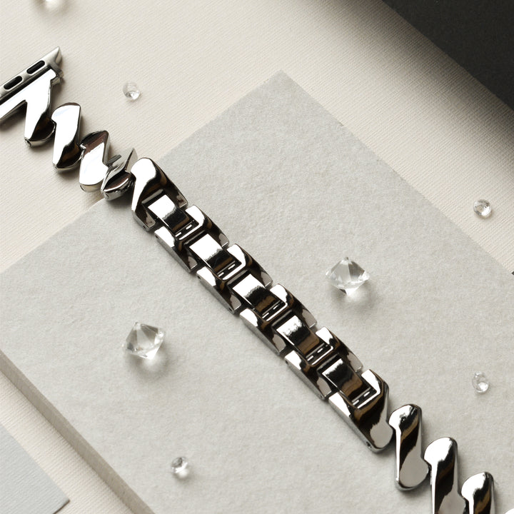 A close-up view of Coconut Lane's Get Wavy Apple Watch Strap - Silver, featuring a polished stainless steel band with a rectangular link pattern, displayed on a light gray backdrop. Small, clear crystals are scattered around the Apple Watch strap, adding a touch of elegance to the composition.