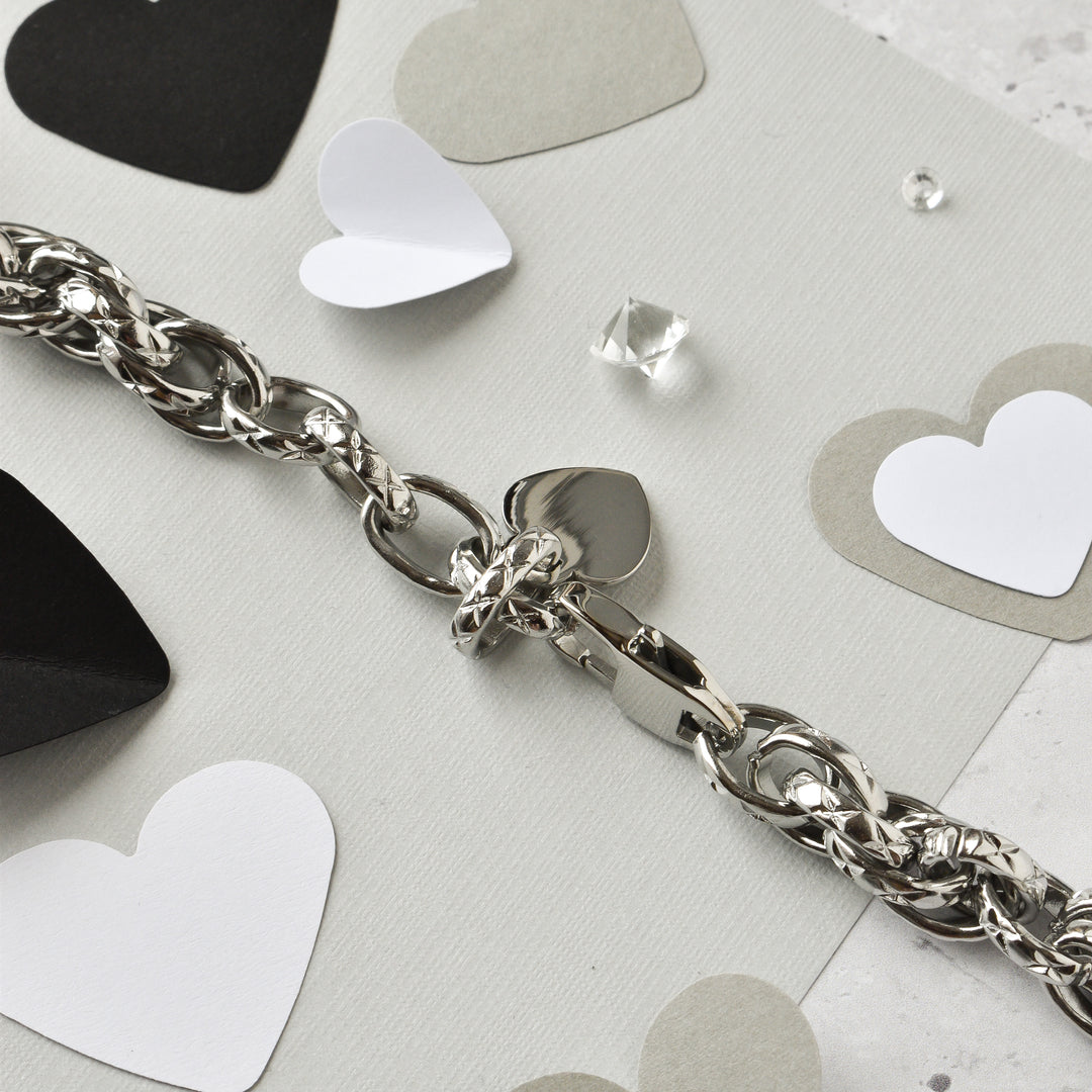 The Chain Bracelet Apple Watch Strap - Silver by Coconut Lane is beautifully displayed on a light gray surface. Surrounding this elegant stainless steel strap are various paper heart cutouts in black, white, and gray, along with a small clear gem, creating a romantic and delicate setting.