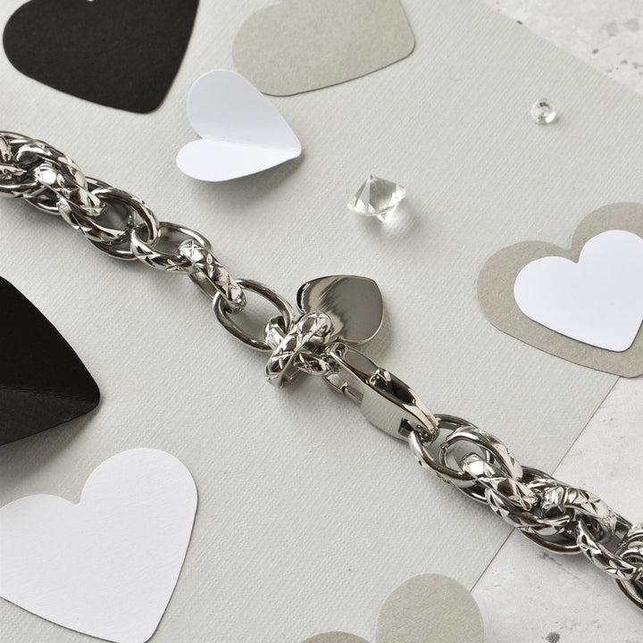 The Chain Bracelet Apple Watch Strap - Silver by Coconut Lane is beautifully displayed on a light gray surface. Surrounding this elegant stainless steel strap are various paper heart cutouts in black, white, and gray, along with a small clear gem, creating a romantic and delicate setting.