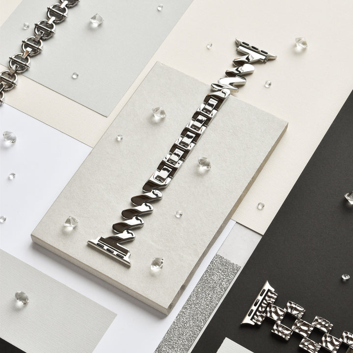 An arrangement of various silver metallic chain links and small, loose diamond-like stones is displayed neatly on overlapping sheets of white, beige, and black paper. At the center is the Get Wavy Apple Watch Strap - Silver by Coconut Lane, twisted and positioned vertically against a neutral background.