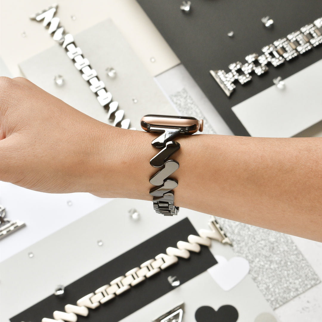 A hand wearing the Get Wavy Apple Watch Strap in black from Coconut Lane, featuring a stylish metallic, zigzag-patterned stainless steel band. Various other watch bands with different designs are displayed in the background on a flat surface, against a backdrop of black, white, and gray colors.