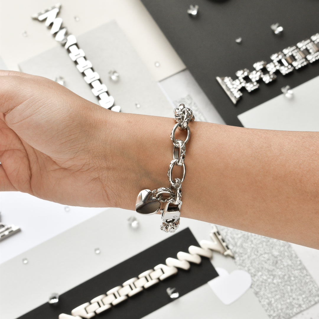 A person's wrist adorned with the Coconut Lane Chain Bracelet Apple Watch Strap in stainless steel, silver, featuring a heart-shaped charm. The background showcases an assortment of silver jewelry on variously textured surfaces, including black, white, and glittery materials.