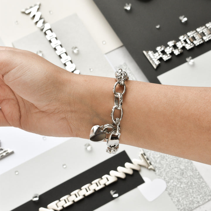 A person's wrist adorned with the Coconut Lane Chain Bracelet Apple Watch Strap in stainless steel, silver, featuring a heart-shaped charm. The background showcases an assortment of silver jewelry on variously textured surfaces, including black, white, and glittery materials.