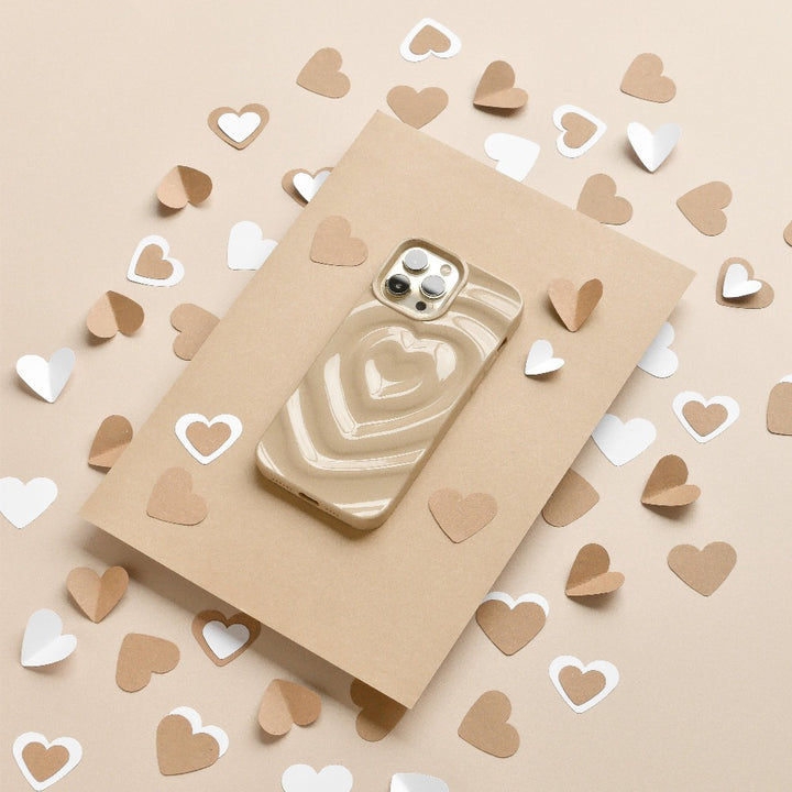 A Coconut Lane Melting Heart Phone Case in Biscuit Beige sits on a brown envelope. The setup is adorned with brown and white paper hearts scattered around, all set against a beige background, creating the perfect scene for this phone case with a raised 3D heart design.