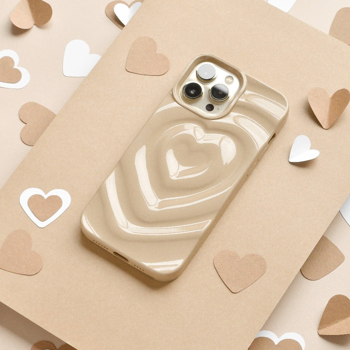 A biscuit beige iPhone with a glossy, extra protective Melting Heart Phone Case by Coconut Lane, featuring a 3D heart design, is placed on a beige surface. The background is decorated with scattered white and brown heart-shaped paper cutouts.