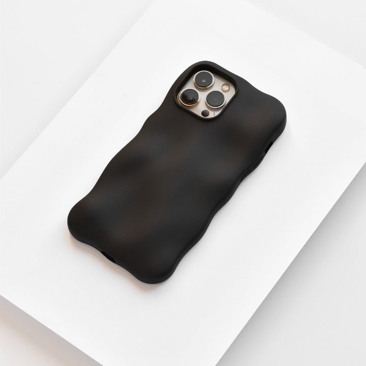 A black iPhone with a Coconut Lane 3D Boujee Phone Case in Matte Black rests on a white surface. The phone's camera array is visible on the back, featuring three lenses and a flash. The matte black finish of the extra protective, minimalist design emphasizes the soft, wavy texture and shape of the case.